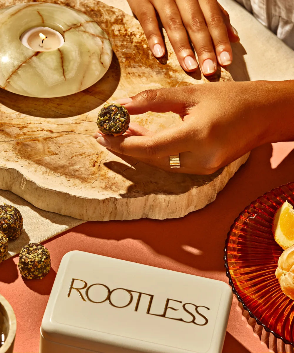 A hand holding an Orange Pistachio Daily Bite beside a Rootless tin, citrus and a candle